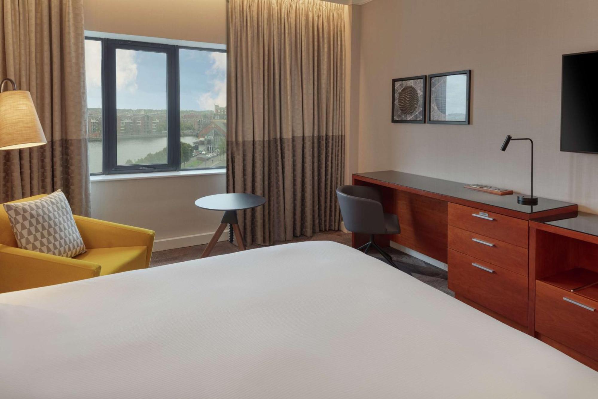 โรงแรมฮิลตัน เบลฟาสท์ เบลฟาสต์ ภายนอก รูปภาพ The photo shows a modern hotel room. It features a large bed with a plain white bedspread, and there's a stylish yellow armchair next to it. A small round table is placed nearby, along with a gray chair. The room has large windows covered with light 