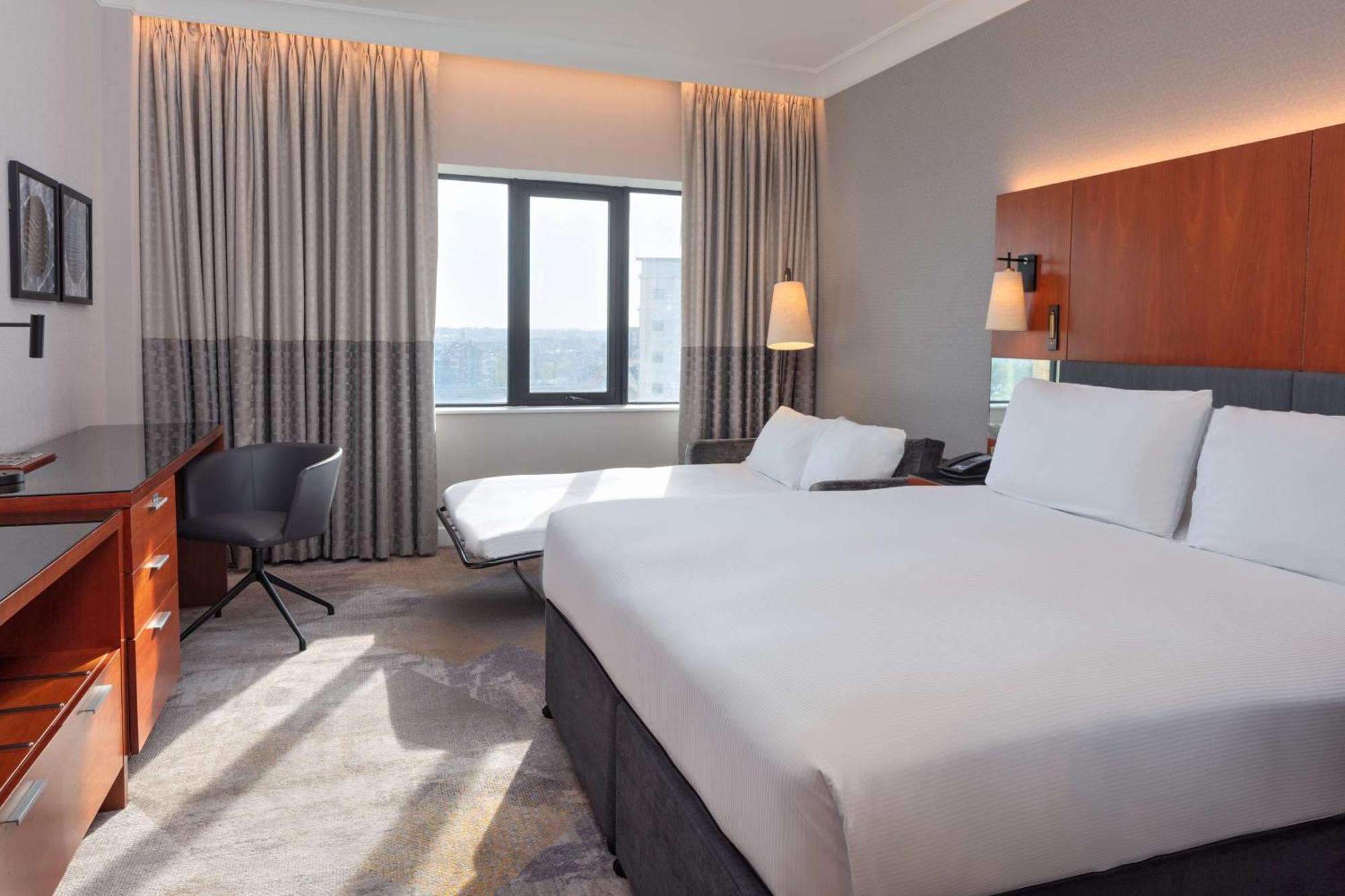 โรงแรมฮิลตัน เบลฟาสท์ เบลฟาสต์ ภายนอก รูปภาพ The photo shows a modern hotel room. It features two beds with white linens and a simple, elegant design. There is a large window allowing natural light to enter, covered by light-colored curtains. A comfortable chair and a small desk are placed near